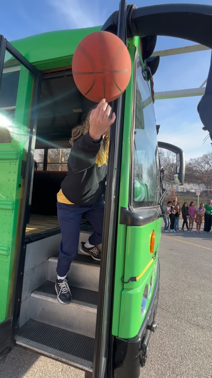 A person steps down from a green bus while spinning a basketball on a fingertip in an outdoor parking area