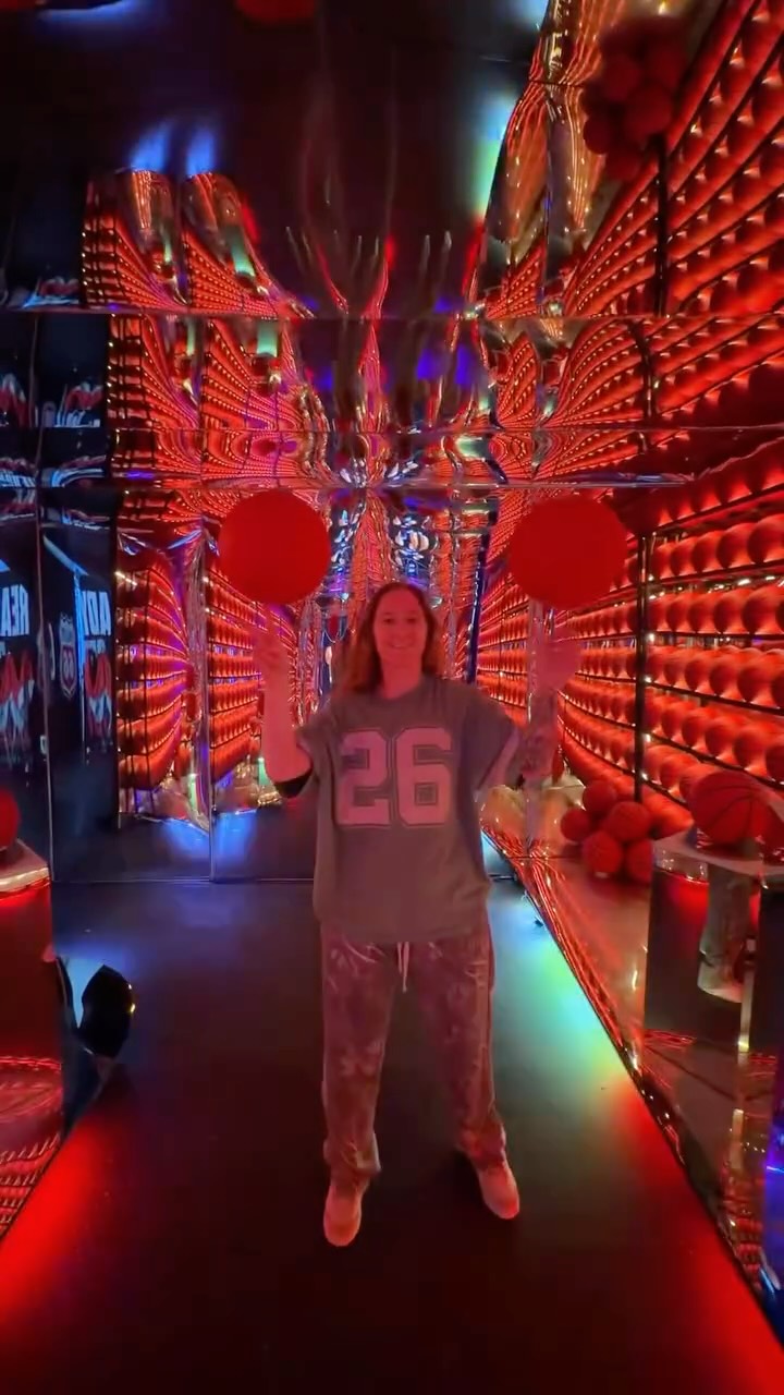 A player in a gray shirt with number 26 spins two basketballs on her fingers in a red-lit hallway lined with basketballs