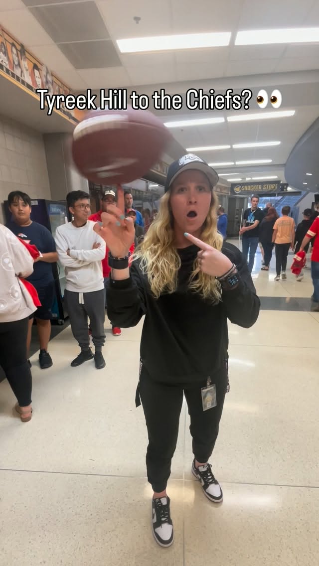 A woman in a black sweatshirt and cap spins an American football on her index finger in an indoor hallway